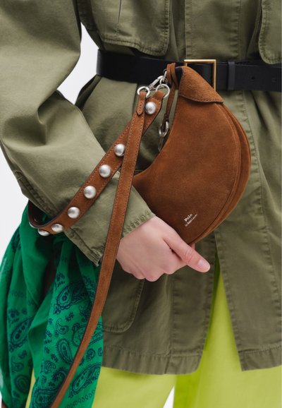 Person holding a brown suede crescent-shaped handbag with silver studs on strap, wearing olive green jacket and lime green pants.
