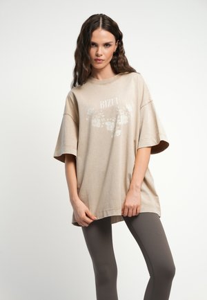 Jonge vrouw met golvend haar die een loszittend beige T-shirt draagt met een bloemenprint en tekst, gecombineerd met aansluitende donkergrijze legging.