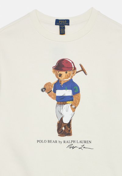 Camiseta de algodón color crema con un gráfico de un oso con camisa polo azul, pantalones blancos y botas marrones, sosteniendo un mazo de polo.