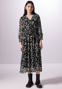 Zwart-witte jurk met patroon, lange lengte, V-hals, gedrapeerde taille en gelaagde rok, gecombineerd met zwarte enkellaarsjes. Doorschijnende stof.