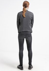 Jeans neri slim-fit abbinati a un blazer scuro, con tessuto strutturato e tasche posteriori distintive, indossati con scarpe nere.