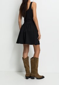 Robe noire sans manches avec une jupe évasée ; modèle portant des bottes de cowboy en daim marron clair avec des clous argentés et des détails noirs au talon.