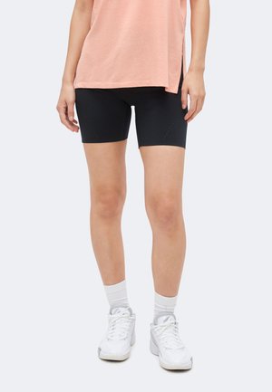 Femeie purtând pantaloni scurți de ciclism negri mulați, top roz lejer, șosete albe și adidași albi sportivă, stând pe un fundal simplu.