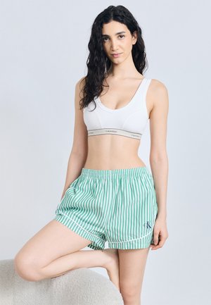 Žena s dlhými kučeravými vlasmi, oblečená v bielom športovom podprsenke a krátkych šortkách so zelenými prúžkami, stojaca s jednou nohou opretou o stoličku.