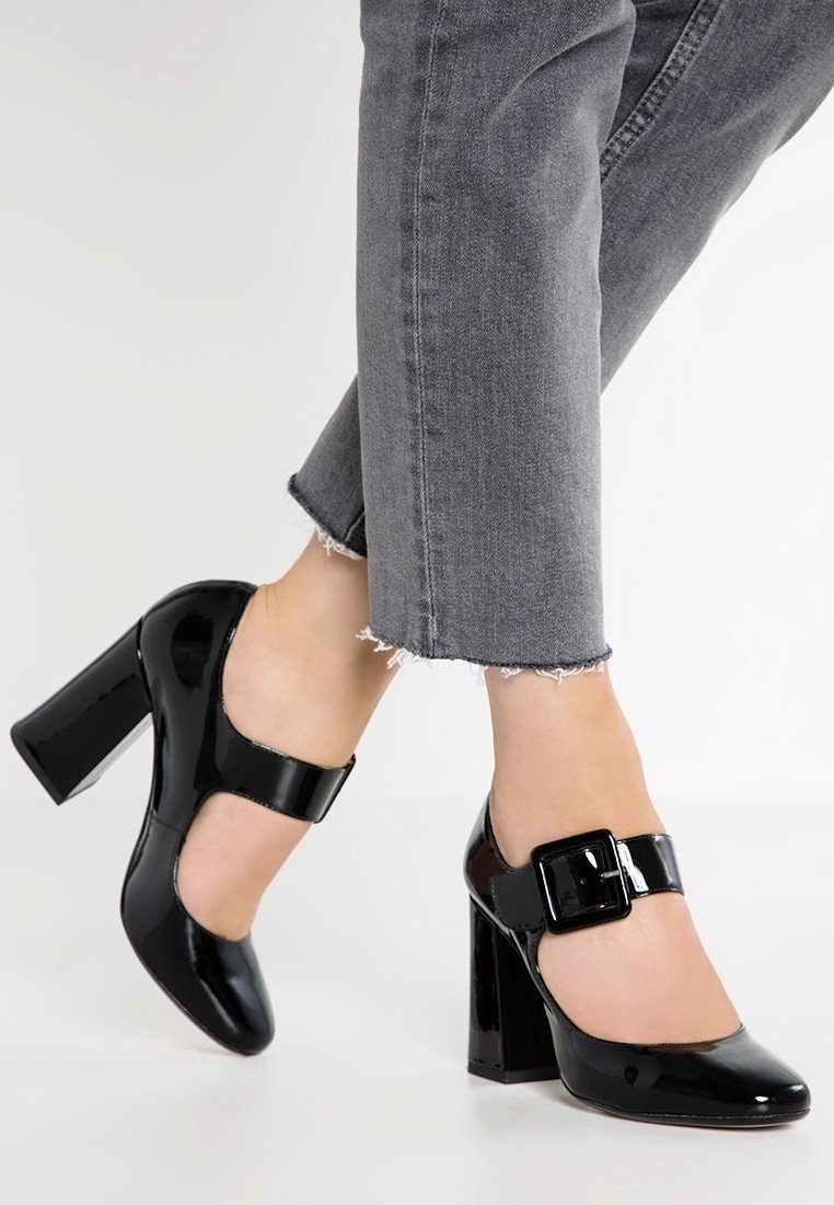 Pieds portant des Mary Jane en cuir verni noir à talons blocs avec de grandes boucles, associées à un jean gris à ourlet effiloché.
