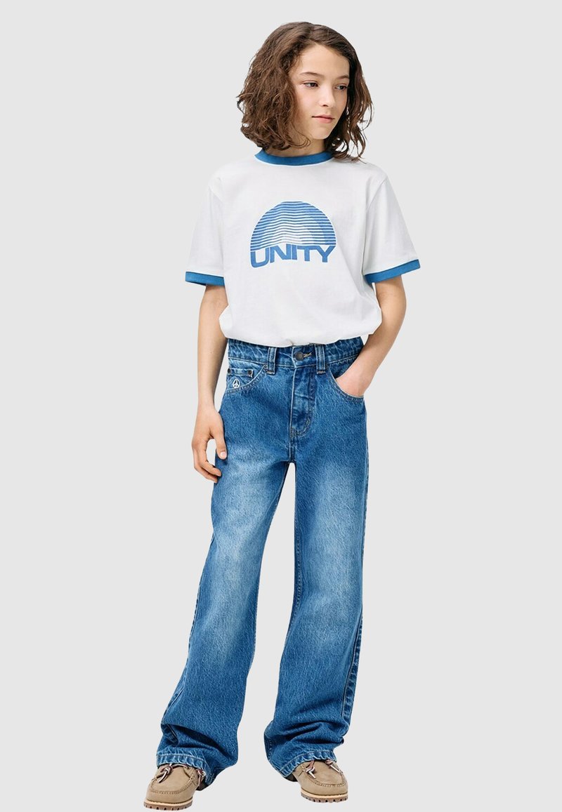 T-shirt blanc avec col contrastant bleu et motif "UNITY". Jeans bleu clair à jambes larges. Chaussures marron clair avec lacets.