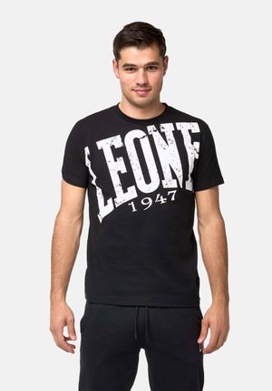 Hombre de pie, mirando al frente, lleva una camiseta negra con un gran texto blanco que dice «"LEONE 1947,"» y pantalones negros a juego.
