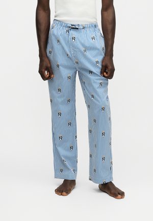 Homme portant un pantalon de pyjama rayé bleu et blanc avec de petits motifs d’ours, debout pieds nus devant un fond blanc uni.
