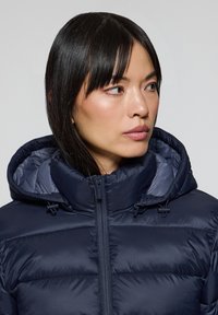 Marinblå pufferjacka med hög krage, huva och dragkedja. Har quiltad struktur med horisontell sömnad.