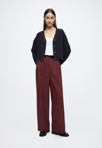 Cardigã preto curto com botões, combinado com calças boca larga em bordeaux de cintura alta. A modelo veste uma camisola de alças branca e sapatos pretos.