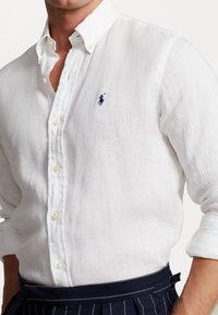 Biała lniana koszula z kołnierzykiem na guziki, przezroczystą plisą na guziki oraz małym niebieskim haftowanym logo na piersi.