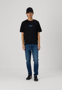 Must arvane t-särk sinise tekstiga, lühikesed varrukad, koos siniste denim teksade ja mustade tossudega, mida täiendatakse musta mütsiga.