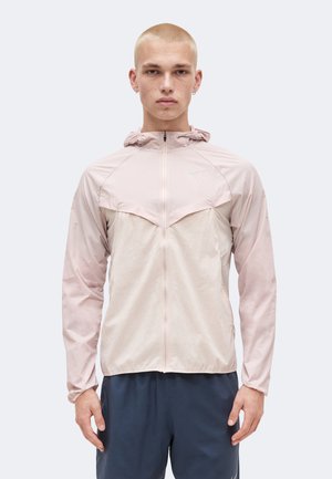 Młody mężczyzna z krótkimi włosami, ubrany w jasnoróżową bluzę z kapturem i zamkiem oraz ciemne spodnie sportowe na jednolitym tle.