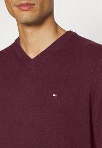 Bordo sweter z dekoltem w serek, wykonany z dzianiny. Posiada małe haftowane logo w czerwonym, białym i niebieskim kolorze na lewej piersi. Gładka faktura.