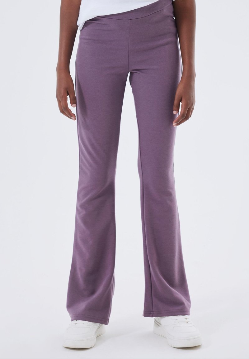 Personne portant un pantalon évasé violet taille haute et des baskets blanches, debout devant un fond blanc uni.