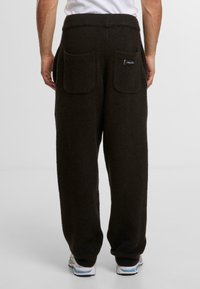 Pantalones de chándal de felpa marrón con una cinturilla de punto. Presenta dos bolsillos de parche cuadrados en la parte posterior y una pequeña etiqueta de la marca en el centro.