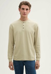 Beżowa, długorękawowa koszula typu henley o teksturowanym wykończeniu, z trzema ciemnymi guzikami przy kołnierzy i ściągaczami na rękawach, zestawiona z niebieskimi dżinsami.