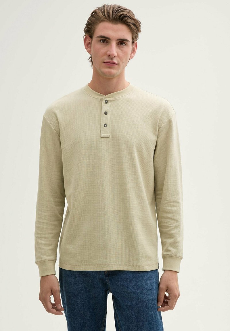 Beżowa, długorękawowa koszula typu henley o teksturowanym wykończeniu, z trzema ciemnymi guzikami przy kołnierzy i ściągaczami na rękawach, zestawiona z niebieskimi dżinsami.