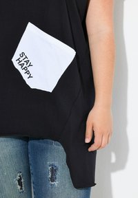 Zwarte top met een witte pocket waarop de tekst "STAY HAPPY" staat. De pocket is gekanteld, wat een ontwerpdetail toevoegt aan de casual look.