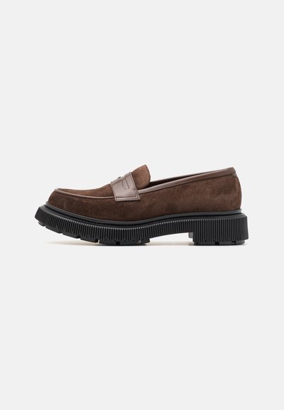 Brūni zamša loafers ar veidotu melnu gumijas zoli, ar dekoratīvu joslu pāri pēdas vidum un izšūtiem akcentiem gar malām.