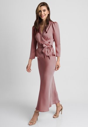 Kobieta uśmiechająca się, ubrana w pudroworóżową satynową bluzkę z kopertowym dekoltem i kokardą oraz dopasowaną długą spódnicę, beżowe sandały na wysokim obcasie, stojąca na białym tle.