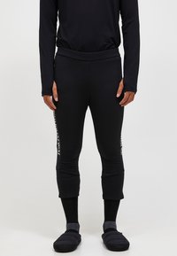 Schwarze sportliche Leggings mit figurbetontem Design und verkürzter Länge, mit weißem Markenlogo an der Seite. Kombiniert mit schwarzen Socken und Hausschuhen.