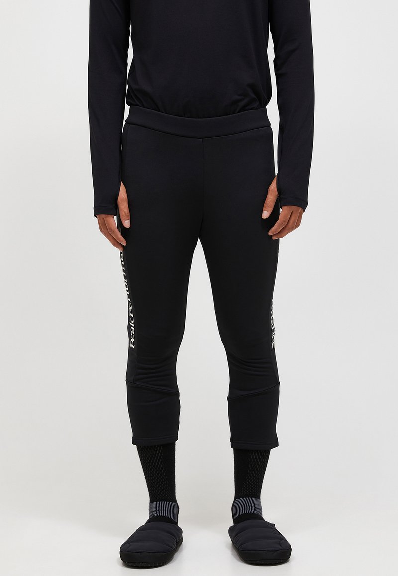 Schwarze sportliche Leggings mit figurbetontem Design und verkürzter Länge, mit weißem Markenlogo an der Seite. Kombiniert mit schwarzen Socken und Hausschuhen.