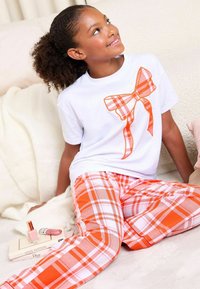 Maglietta bianca con grafica di fiocco a quadri arancioni, abbinata a pantaloni a quadri arancioni e bianchi. Tessuto morbido, design casual, vestibilità rilassata.