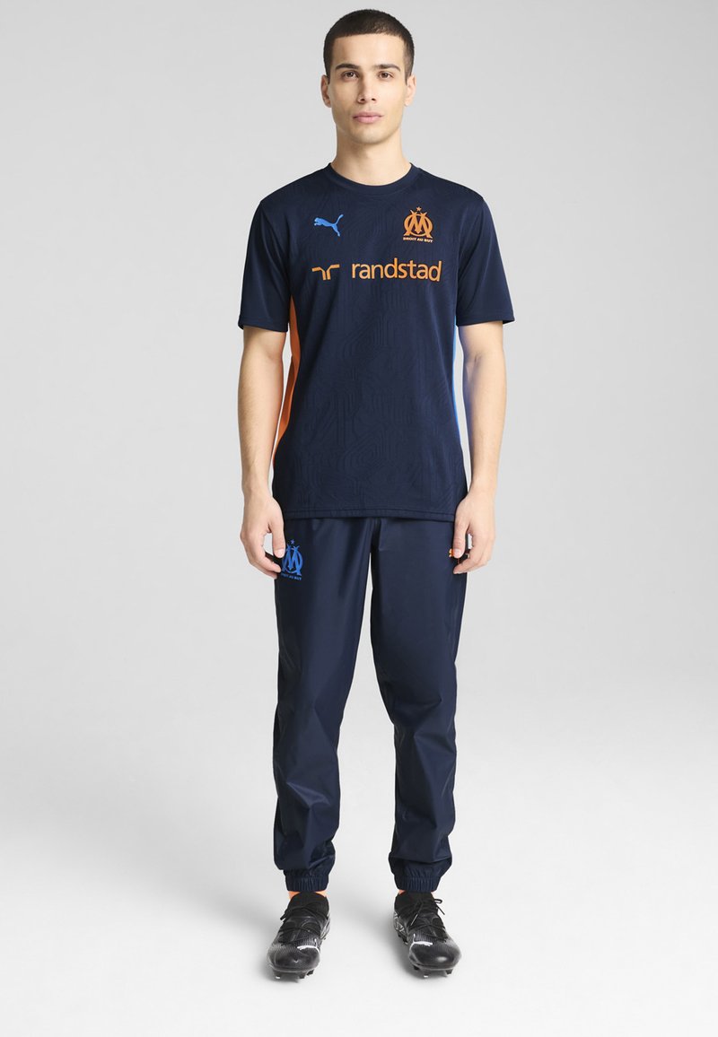 T-shirt de sport marine avec des accents et un logo orange, associé à un pantalon marine. Design texturé pour le t-shirt ; le pantalon est doté de poignets élastiques et d'un cordon de serrage.
