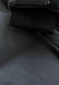 Veste puffer noire avec une texture lisse et un éclat subtil. L'étiquette présente le mot "BAKER" dans une finition ton sur ton. Détail en gros plan du tissu.