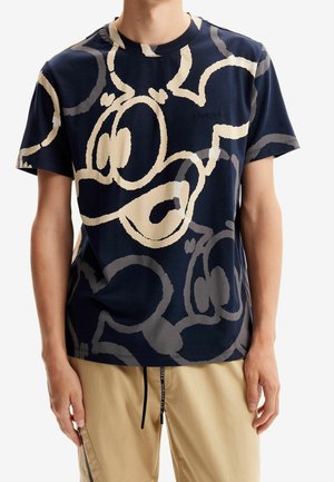 Homme portant un t-shirt bleu marine avec de grands motifs abstraits de visages de dessin animé crème et gris et un pantalon beige avec un cordon noir.