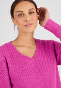 Maglione rosa a maglia con scollatura a V, caratterizzato da un motivo testurizzato nella parte superiore e polsini a coste. Materiale morbido, vestibilità casual.