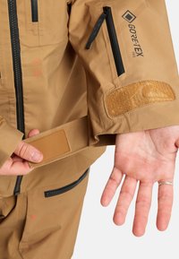 Kurtka GORE-TEX PRO w kolorze brązowym o teksturowanym wykończeniu, z czarnym zamkiem błyskawicznym, regulacją mankietów na rzep oraz widocznymi detalami z logo.