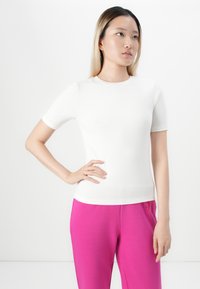 Ungdom med rett blondt hår har på seg en hvit t-skjorte med korte ermer og lyse rosa bukser, poserer med den ene handen på hoften.