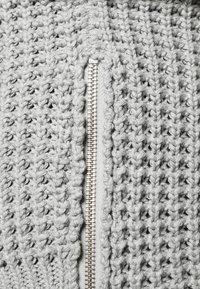 Gros plan sur une fermeture éclair en métal argenté sur un tissu tricoté épais gris, montrant la texture détaillée du tricot et les dents de la fermeture éclair.