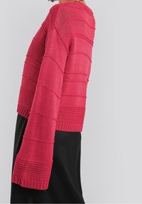Kurzer rosa gestrickter Pullover mit einem cropped Design, der strukturierte horizontale Streifen und weite Ärmel aufweist. Schwarzer Stoff darunter.