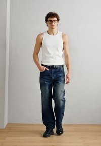 Débardeur blanc à côtes, jeans bleu foncé décolorés, ceinture noire et baskets noires. Le modèle se tient sur un sol en bois, avec un fond clair et uni.