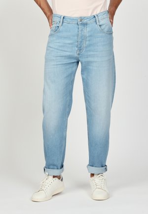 Mann trägt hellblaue, umgekrempelte Jeans und weiße Sneakers und steht mit den Händen in den Taschen vor einem einfarbigen Hintergrund.