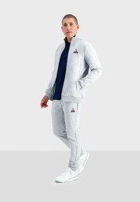 Completo sportivo grigio composto da giacca con zip e pantaloni aderenti, realizzato in tessuto morbido, con logo sul petto e sulla coscia. Sneakers bianche.