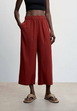 Person iført højtaljede, vidde bukser i rustfarve, sort crop top og beige sandaler med åbne tåspidser, stående på en lys gulv.