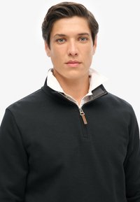 Pullover nero con zip e finitura testurizzata, dotato di chiusura con zip e un colletto bianco a contrasto sotto.
