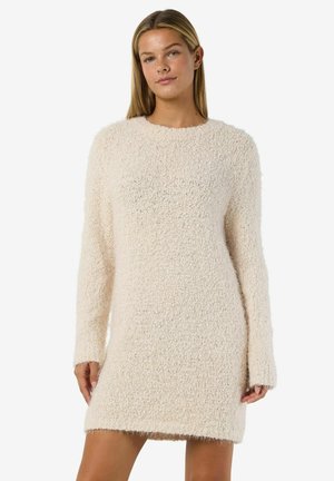 Ung kvinna som bär en långärmad, strukturerad beige stickad klänning, stående framför en enkel vit bakgrund.
