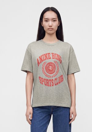 Femme aux longs cheveux foncés portant un t-shirt gris Anine Bing Sports Club et un jean bleu, debout devant un fond clair uni.