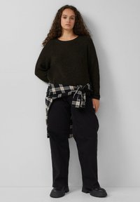 Schwarzer Strickpullover, lockere Passform, kombiniert mit schwarzen weiten Hosen. Karohemd in den Farben Grün, Creme und Blau um die Taille gebunden. Schwarze Stiefel.
