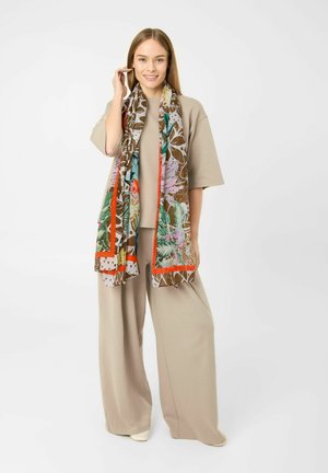 Frau trägt ein beiges Oversize-Outfit und einen langen bunten Blumenschal, steht vor weißem Hintergrund und lächelt.