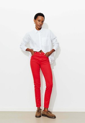 Personne debout contre un mur blanc, portant une chemise blanche boutonnée, un pantalon rouge et des chaussures à imprimé léopard, les mains posées sur les hanches.