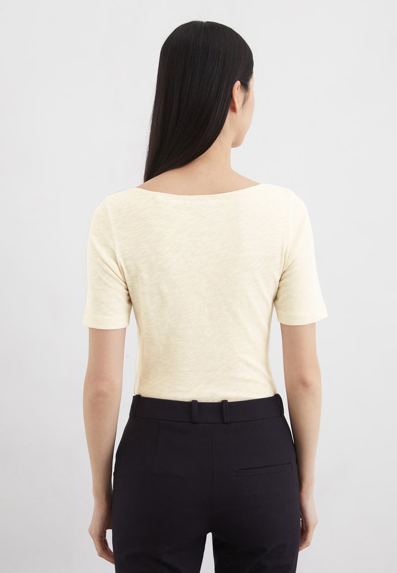 Helles gelbes Kurzarmoberteil mit Rundhalsausschnitt, kombiniert mit dunklen Hosen. Der Stoff wirkt strukturiert und lässig.