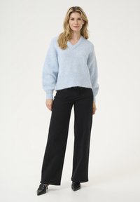 Helles Blau, übergroßer V-Ausschnitt-Pullover aus weichem, strukturiertem Strick mit geripptem Saum. Kombiniert mit hochgeschnittenen schwarzen weiten Jeans und spitzen schwarzen Schuhen.