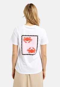 Weißes T-Shirt mit einem rosa rechteckigen Grafikdruck, der zwei orangefarbene Krabben und den Text "JA, MEER" zeigt, eingerahmt von einem schwarzen gewellten Rand.