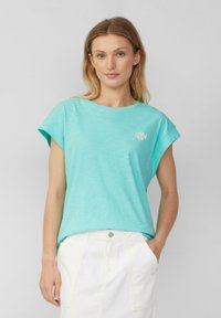 Vrouw met blond haar, gekleed in een turkooizen t-shirt met korte mouwen en een klein vislogo, in combinatie met een witte rok, tegen een effen achtergrond.